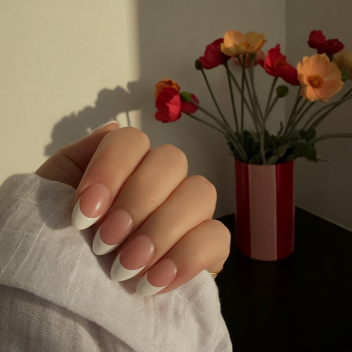 A close-up of a hand with a classic French manicure, by instant Mani Co. press on nails, resting on a white textured fabric sleeve. In the softly lit background, a red and pink striped vase holds a bouquet of red and orange flowers.