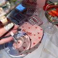 Girl wearing sparkly nails. holding champaign, in front of pink heart cake, with Lover Instant Mani Co press on nails in background.
