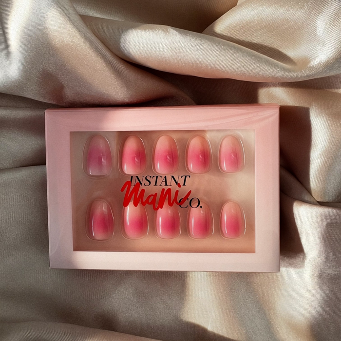 A set of 10 pink press-on nails with a gradient design, displayed in a light pink box with the brand name 'Instant Mani Co.' printed on the front, set against a draped champagne-colored satin fabric.
