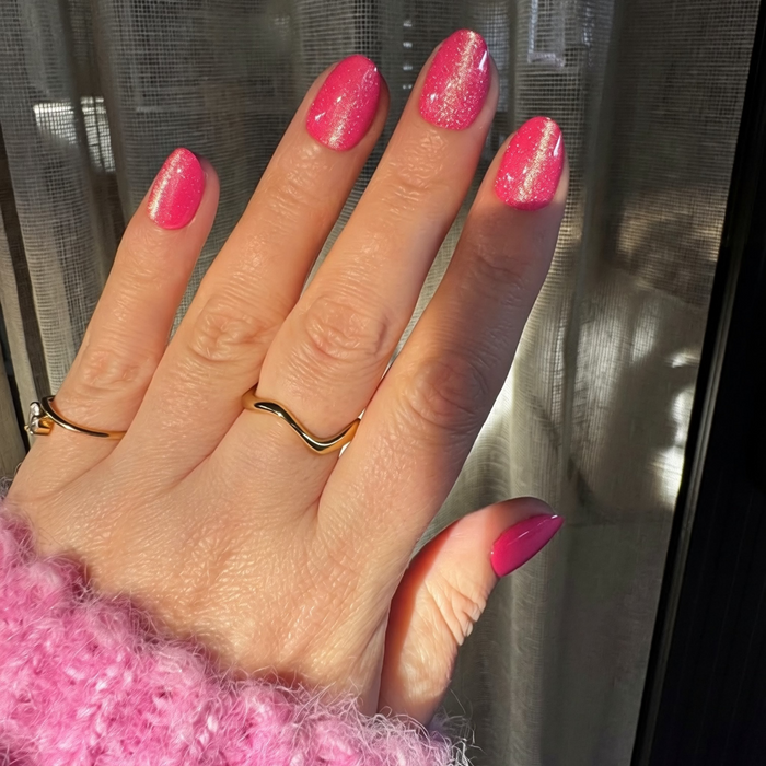A close-up of a hand with bright velvet Instant Mani Co. press on nails, wearing gold rings and a fuzzy pink sweater, with sheer curtains in the background.
