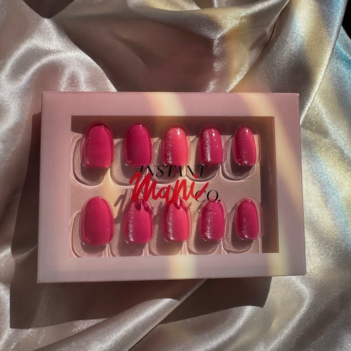 A set of Instant Mani Co Velvet pink press-on nails in a light pink box with the brand name 'Instant Mani Co.' printed on the front, styled on a shiny champagne satin fabric background.