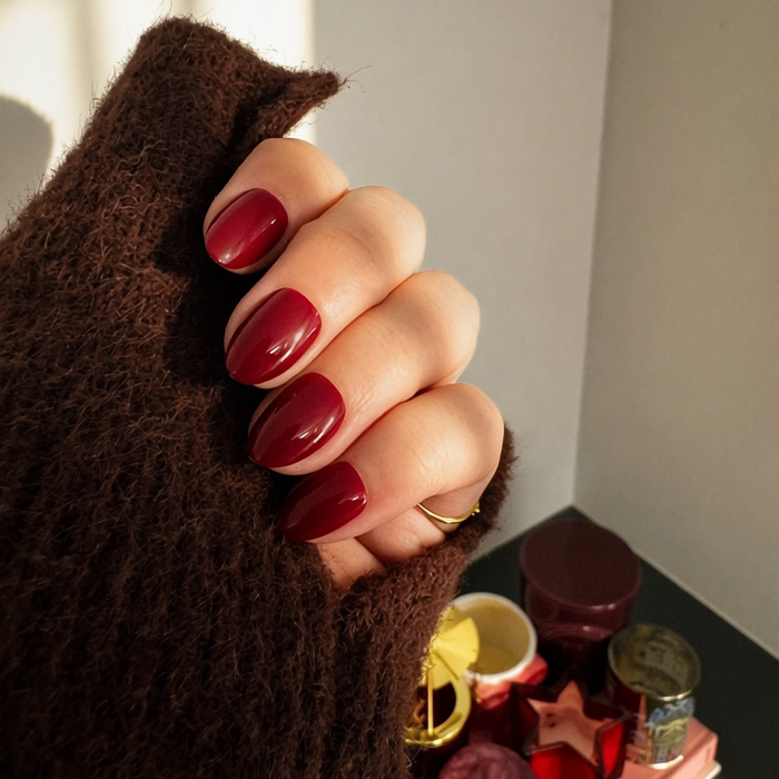 A close-up of a hand with glossy Instant Mani Co. burgundy Merlot press on nails, wearing a brown long sleeve jumper, with candles in the background