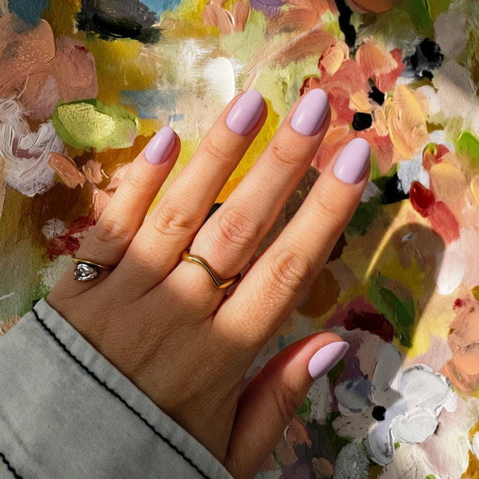A close-up of a hand with Instant Mani Co. Lilac Haze press on nails resting on a colorful, textured oil painting palette. The hand is wearing a gold ring and a thin gold band.