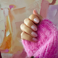 A close-up of a hand wearing glossy pink Instant Mani Co Press on nails in Strawberry Glaze resting on a fluffy bright pink knit sleeve, set against an abstract pastel painted background.