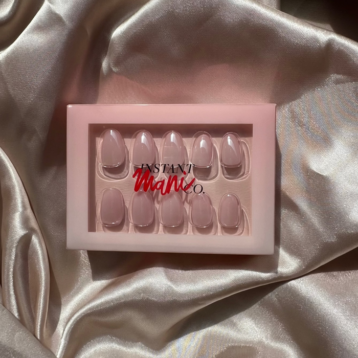 A set of 10 press-on nails in a pale pink shade, presented in a light pink box with the brand name 'Instant Mani co' visible, set against a draped champagne satin fabric background.