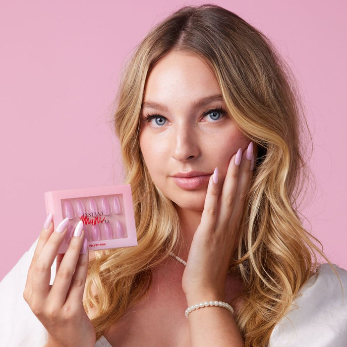 Girl wearing Instant Mani Co Velvet Pink Press on nails standing in front of pink background wearing white shirt